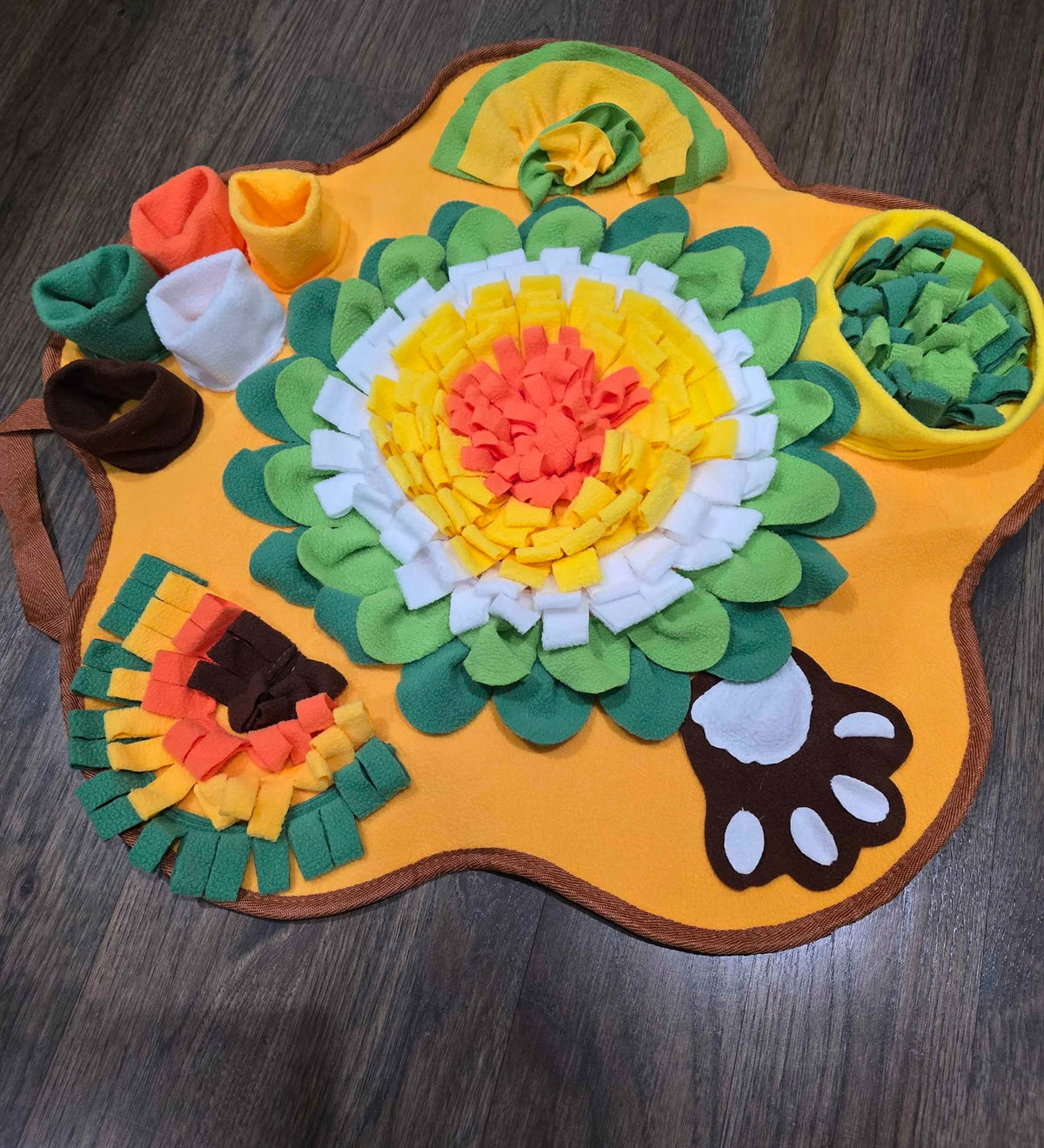 A yellow flower-shaped snuffle mat on a wood floor, its fleece petals and pockets hiding kibble for a dog to sniff out.
